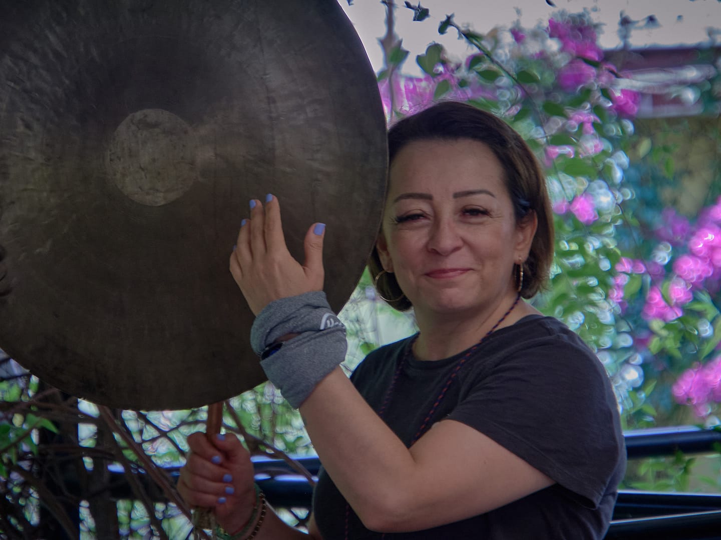 Terapeuta de sonido de Eco Ancestral sonriendo mientras sostiene un gong terapéutico en un entorno natural con fondo de flores. Sesión de sonoterapia al aire libre
