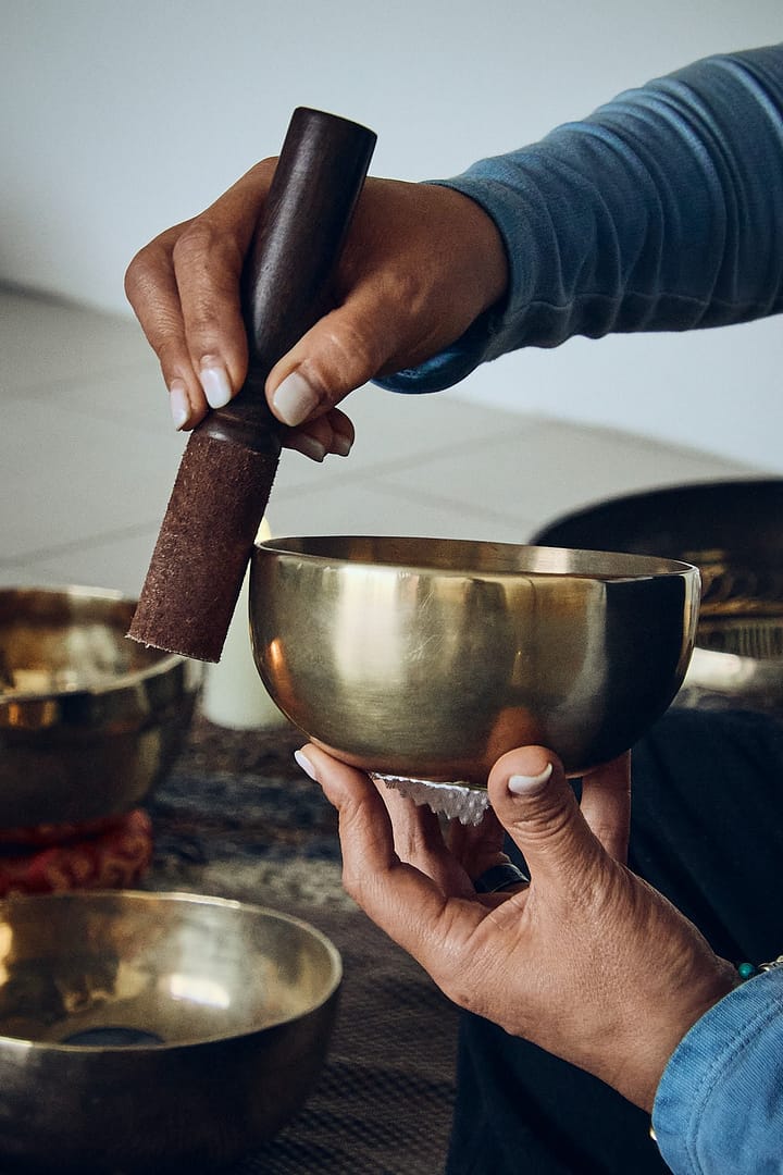 Primer plano de manos sosteniendo y tocando un cuenco tibetano de latón con un mazo de ante, mostrando la técnica de sonoterapia.