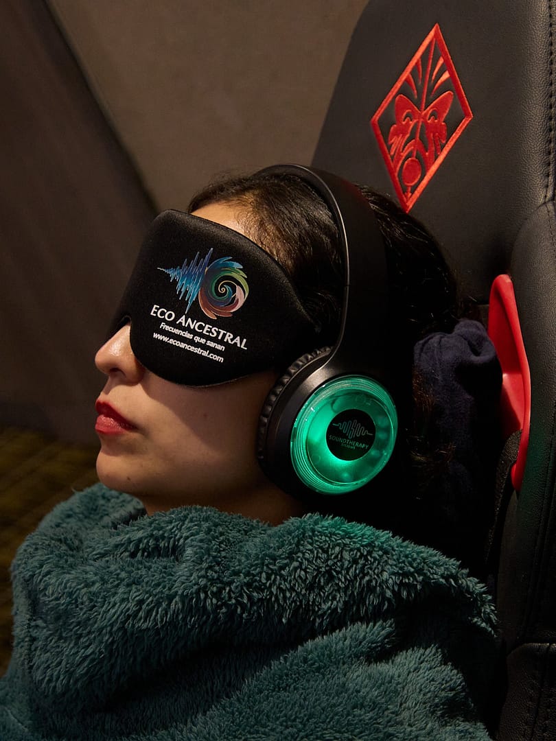 Mujer en estado de relajación profunda utilizando auriculares iluminados de terapia de sonido y antifaz corporativo de Eco Ancestral durante una sesión inmersiva.
